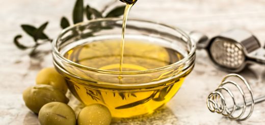 Close-up of olive oil being poured into a glass bowl surrounded by fresh olives and kitchen tools.